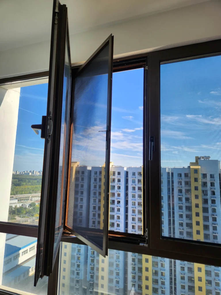 Dark aluminum casement window opened outward with a built-in high-visibility fly screen facing city buildings.