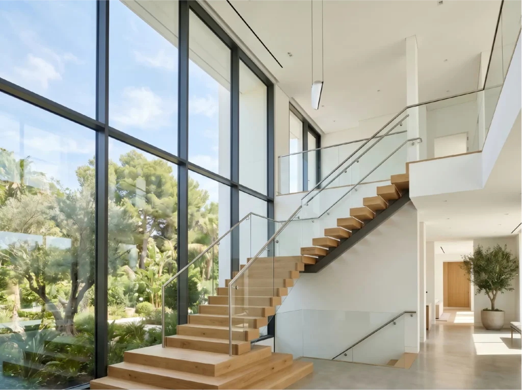 A dramatic two-story staircase in a modern luxury home featuring a massive, fixed structural window wall overlooking a tropical garden.
