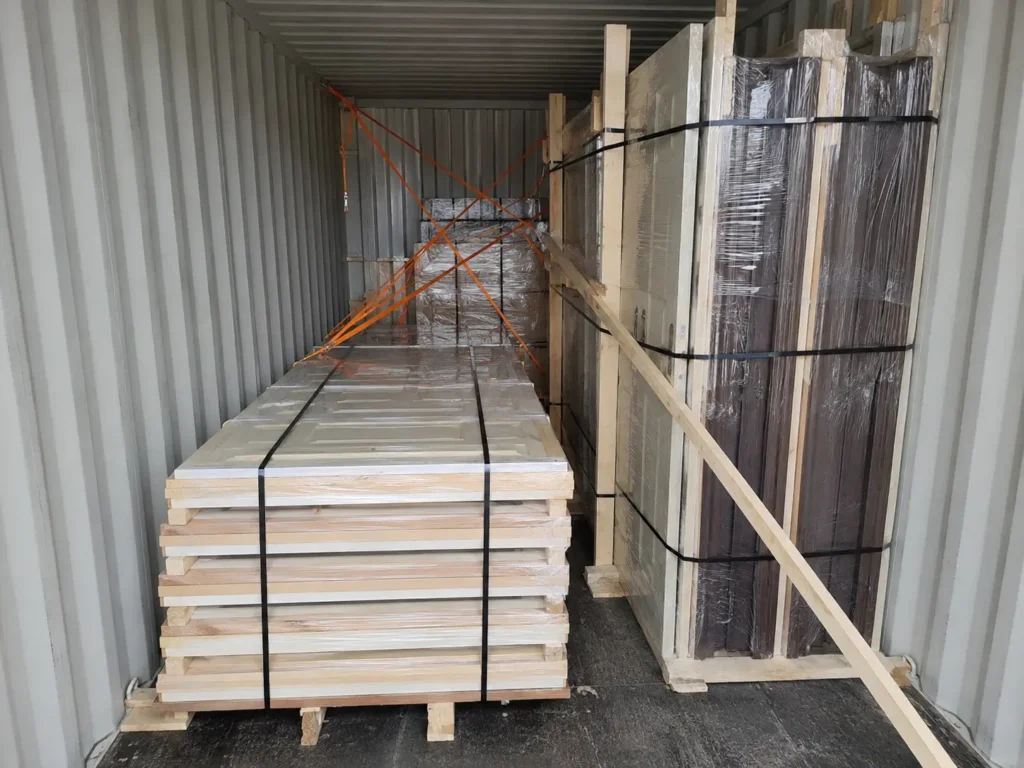 Wooden doors loaded inside a shipping container with pallets, straps, dunnage, and bracing to prevent cargo shifting.